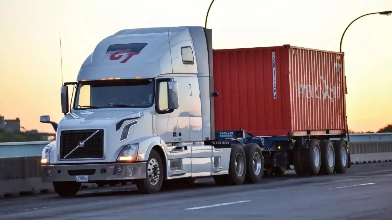 A white GT Group Volvo tractor hauling a red shipping container on a highway overpass at sunset